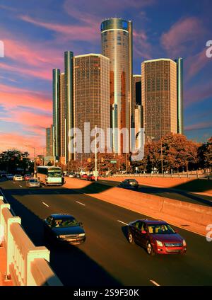 Ein urbanes Stadtbild mit einem hoch aufragenden modernen Gebäudekomplex, der unter einem Abendhimmel mit Orangen- und Rosa-Tönen beleuchtet wird, während Fahrzeuge fahren Stockfoto