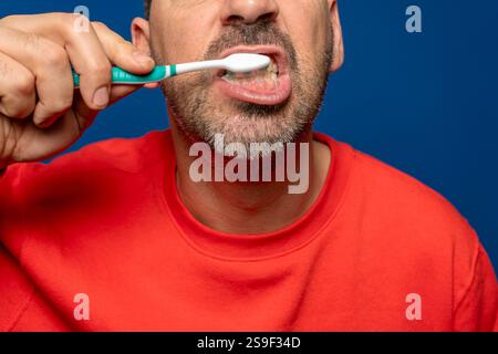 Nahaufnahme eines bärtigen hispanischen Mannes in seinen 40ern, der seine Zähne putzt, isoliert auf blauem Atelierhintergrund. Zahnhygiene und Gesundheitskonzept. Stockfoto