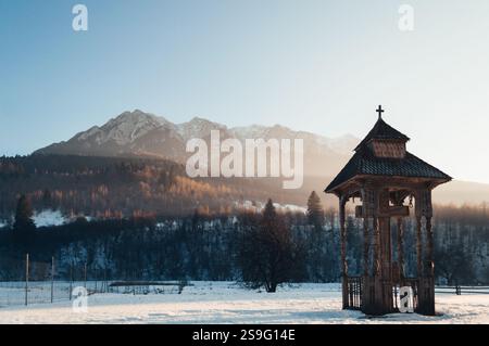 Eine ruhige Winterszene mit einem traditionellen rumänischen Holzpavillon mit Kreuz vor den schneebedeckten Piatra Craiului Bergen. Das golde Stockfoto