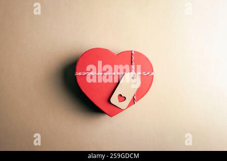 Rote Box in Herzform mit einem Kraftanhänger auf beigefarbenem Hintergrund, das Liebe und nachdenkliches Geschenk darstellt. Valentinstag-Konzept. Draufsicht, flach. Stockfoto