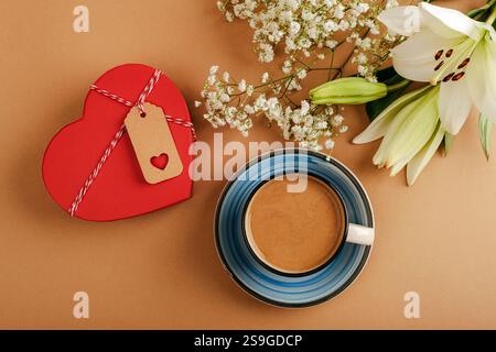 Rote, herzförmige Box mit Blumen und einer Tasse Kaffee auf beigefarbenem Hintergrund, symbolisiert Liebe und Entspannung. Valentinstag-Konzept. Draufsicht, flach Stockfoto