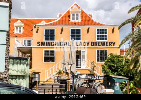 Die Fassade der Swinging Old Lady Brewery in der Breedestraat in Otrabanda, Willemstad, Curacao. Das gelbe Kolonialgebäude mit seiner Niederländisch-karibischen Giebelarchitektur und einer Außentreppe beherbergt die Brauerei, die ihren Namen vom umgangssprachlichen Namen für die Queen Emma Pontoon Bridge ableitet; Bierfässer vor dem Eingang weisen darauf hin, dass das Gebäude als Brauerei genutzt wird. Die Swinging Old Lady Brewery in Willemstad, Curacao, ist nach der berühmten Pontonbrücke benannt Stockfoto