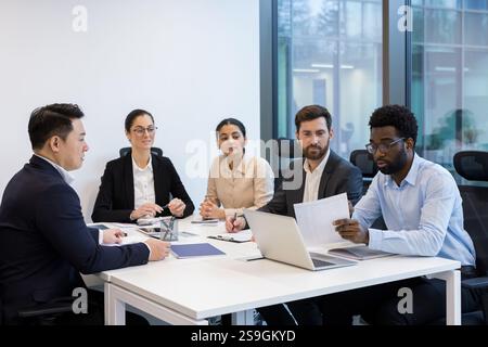 Büroangestellte, in Business-Anzügen, die am Schreibtisch sitzen, sich unterhalten und Details über neue Projekte und Startup diskutieren, erfolgreiche Geschäftsleute sind vielfältig. Geschäftsleute diskutieren elektronische Berichte. Stockfoto