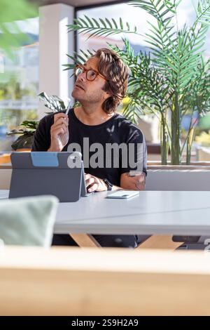Ein Mann sitzt an einem Tisch mit einer Tablette und einem Stift. Er schaut an die Decke und denkt nach. Stockfoto