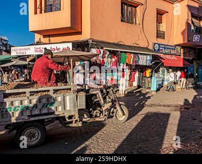 Geschäftiger Marktplatz in Marrakesch mit farbenfrohen Bekleidungsständen und Einkaufsmöglichkeiten, während ein Motorradwagen durch die Kopfsteinpflasterstraße fährt. Stockfoto
