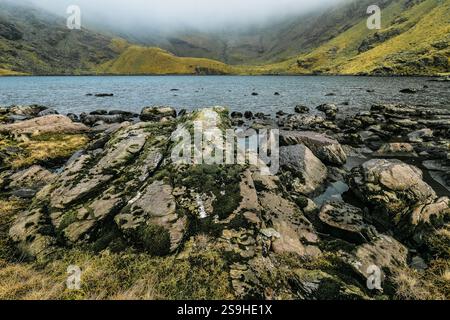 Ein Schuss eines zerklüfteten Berges, bedeckt von grüner Vegetation und umhüllt von Nebel. Am Fuße des Berges befindet sich ein See, dessen Oberfläche die c Stockfoto
