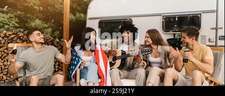 Interrassische Freunde, die Gitarre spielen und singen, amerikanische Flagge halten, Bier trinken, in den Sommerferien in der Nähe von RV sitzen Stockfoto