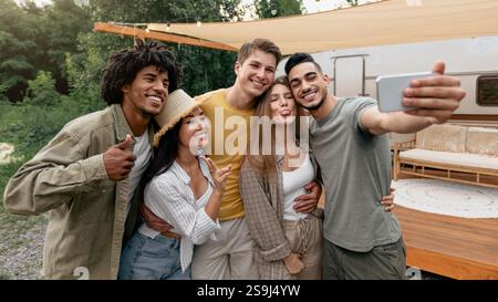 Glückliche, vielfältige Freunde posieren in der Nähe von Wohnmobil auf dem Land, machen verschiedene Gesten, Selfie auf dem Smartphone Stockfoto