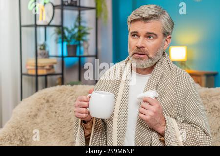 Kranker müder reifer Mann, der auf dem Heimsofa sitzt, bläst niesst Tücher Rotz in Serviette Taschentuch, trinkt heißes Getränk oder nimmt Medikamente aus dem Becher. Der Typ scheint kalt, Schwäche, Unwohlsein, laufende Nase, Krankheit Stockfoto