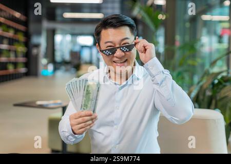 Glücklicher, reifer chinesischer Geschäftsmann, der eine Sonnenbrille mit Geldgeld-Geldscheine in der modernen Bürolobby trägt. Erfolg Geschäftslaufbahn, Lottosieger, großes Einkommen, Reichtum. Stockfoto