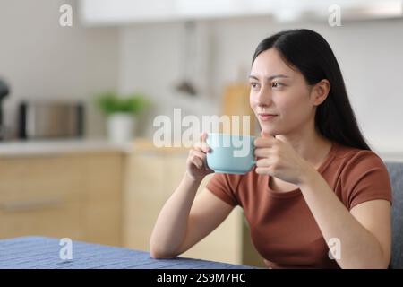 Entspannte asiatische Frau, die zu Hause in der Küche trinkt und nachdenkt Stockfoto