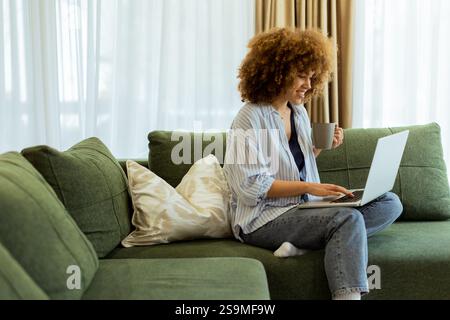 Eine junge Frau sitzt bequem auf einem weichen Sofa und genießt ihren warmen Kaffee, während sie an ihrem Laptop arbeitet, beleuchtet von sanftem Tageslicht in einem stilvollen Ambiente Stockfoto