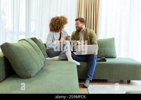 Ein fröhliches Paar sitzt nebeneinander auf einer weichen grünen Couch und führt ein aufregendes Gespräch, während es unter warmem Su auf einem Laptop Ideen durchstöbert Stockfoto