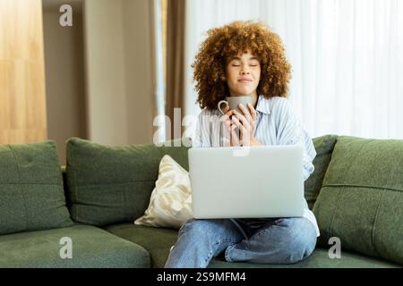 Eine junge Frau sitzt bequem auf einer weichen, grünen Couch und genießt einen Dampfbecher, während sie in einem sonnendurchfluteten Zuhause an ihrem Laptop arbeitet Stockfoto