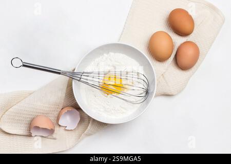 Eigelb und Mehl in einer Schüssel verquirlen. Hühnereier auf beiger Serviette. Salz und eine Flasche Wasser. Weißer Hintergrund. Flache Lagen Stockfoto