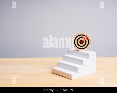 Zielsymbol auf rundem Holzblock auf 3D-weißen Stufen als Balkendiagramm auf Holztisch, weißer Hintergrund mit Kopierraum. Business Growth Plan, A Stockfoto