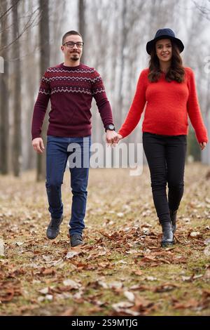 Glücklich erwartendes Paar, das Hand in Hand durch einen Park mit gefallenen Blättern geht, sich gegenseitig anlächelt, mit einem verschwommenen Hintergrund, der ihre Verbindung unterstreicht Stockfoto