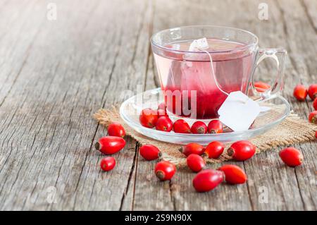 Eine Glasschale Hagebutten-Kräutertee mit Teebeutel und rosafarbenen Früchten vor rustikaler Holzkulisse. Dogrose Winter Tee Hintergrund Stockfoto