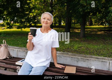 Porträt einer entspannten europäischen grauhaarigen älteren Frau, die auf der Bank sitzt, aus dem Becher schlürft und am Sommertag im öffentlichen Stadtpark im Freien genießt Stockfoto
