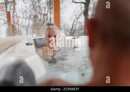 Baden im Wannen-Whirlpool an kalten Wintertagen. Eine ältere Frau sitzt im Wasser und spricht mit dem Ehemann. Stockfoto