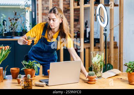 Professionelle junge asiatische Gärtnerin, die Vlog-Session auf Pflanzen Töpfen Arrangement-Kurs mit Laptop und Telefon Kamera zu Hause macht. Selbständige Erwerbstätigkeit Stockfoto