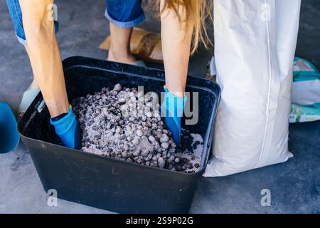 Nicht erkennbare Hausfrau in Handschuhen und lässiger Hauskleidung, die Boden für Pflanzentransplantation in Garage oder Versorgungsraum vorbereiten. Stockfoto