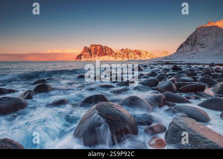 Winterlandschaft mit schneebedeckten Berggipfeln, felsiger Küste und malerischem Sonnenuntergang über Uttakleiv Beach, Lofoten Inseln, Norwegen Sonnenuntergang Stockfoto