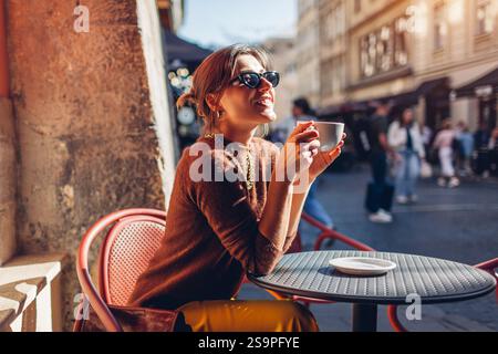Glückliche, stilvolle Frau, die im Café im Freien sitzt und Kaffee trinkt. Frau genießt die Architektur der Stadt Lemberg auf der Straße und entspannt sich auf der Terrasse. Stockfoto