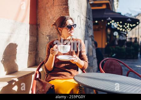 Porträt einer stilvollen Frau, die im Café sitzt und Kaffee trinkt. Frau genießt die Architektur der europäischen Stadt Lviv auf der Straße und entspannt sich auf der Terrasse. Stockfoto