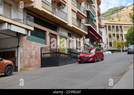 Eine Stadtstraße mit geparkten und fahrenden Autos in der Nähe von Wohngebäuden auf einem Hügel. Die Szene fängt eine lebhafte urbane Umgebung mit dem Alltag und ein Stockfoto