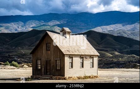 Ein rustikales Holzhaus steht allein in einer kargen Landschaft, umgeben von sanften Hügeln und Bergen unter einem bewölkten Himmel. Die Struktur weist eine Neigung auf Stockfoto