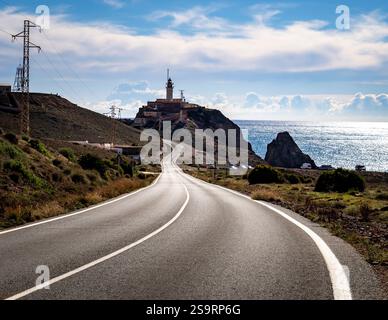Eine gewundene Straße, die zu einem Leuchtturm auf einer Klippe am Meer führt, mit einem klaren blauen Himmel und fernen Wolken. Die Landschaft bietet felsiges Gelände und Stockfoto