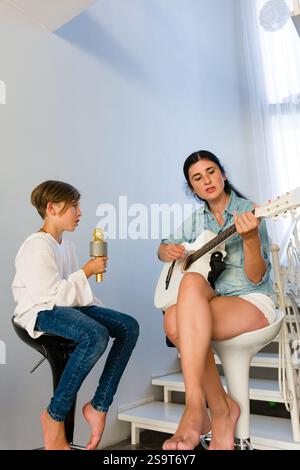 Ein Junge benutzt ein Mikrofon, während seine Mutter eine weiße Akustikgitarre spielt. Sie sitzen auf Hockern nahe einer Treppe in einem hellen Haus. Stockfoto