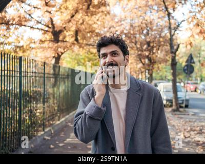 Porträt eines jungen lächelnden Mannes aus dem Nahen Osten, der auf dem Handy spricht und Gespräche führt, während er an einem schönen Herbsttag auf der Straße spaziert. Stockfoto