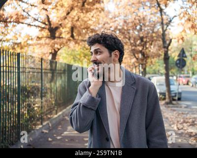 Porträt eines jungen lächelnden Mannes aus dem Nahen Osten, der auf dem Handy spricht und Gespräche führt, während er an einem schönen Herbsttag auf der Straße spaziert. Stockfoto