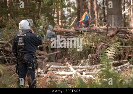 In einer Waldlandschaft steht ein Polizist vor einer provisorischen Barrikade, in der sich Demonstranten versammeln. Die Demonstranten zeigen eine Regenbogenfahne, ich Stockfoto