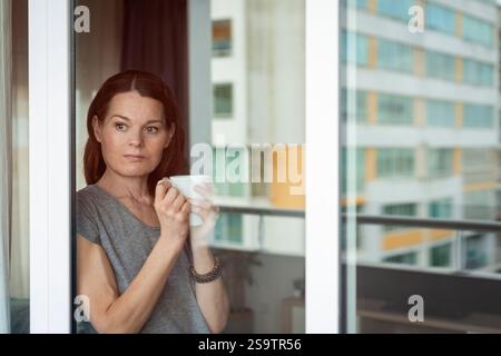 Frau, die an einem Fenster steht und einen Becher hält, Reflexionen Stockfoto