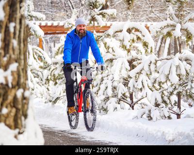 Ein fröhlicher Mann fährt mit dem Fahrrad in einem Winterpark zwischen schneebedeckten Bäumen. Bärtiger Radfahrer in blauer Jacke. Aktiver Lebensstil Stockfoto