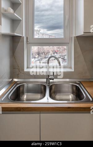 Doppelschüssel Küchenspüle vor dem Fenster. Eine Doppelschüssel aus Edelstahl-Küchenspüle auf einer Holzarbeitsfläche. Stilvolle Küche Waschbecken mit Holzkabine Stockfoto