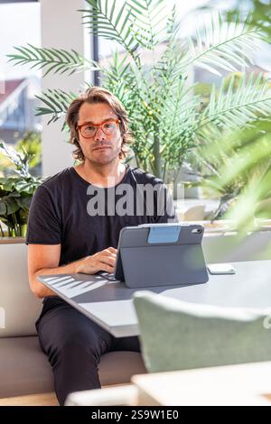 Ein ernster Mann sitzt an einem Tisch und arbeitet mit seinem Tablet, vertikalem Foto. Er trägt ein schwarzes Hemd und eine Brille Stockfoto