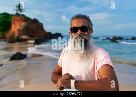 Senior Indianer, grauer Bart, Sonnenbrille am Strand benutzt Smartwatch. Der ältere überprüft den Fitness-Tracker mit kabellosen Ohrstöpseln. Der stilvolle Opa genießt Technik Stockfoto