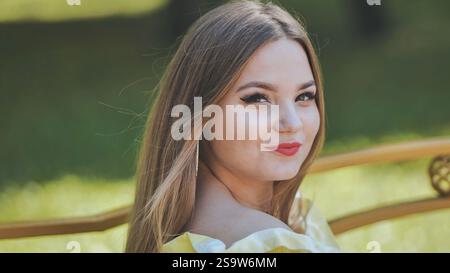 Junge Frau mit langen braunen Haaren, rotem Lippenstift, lächelnd, während sie an sonnigen Sommertagen auf goldenen Parkbank sitzt Stockfoto