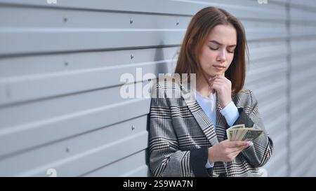 Eine junge Geschäftsfrau, die sich Sorgen macht, während sie ihre letzten Dollar zählt, mit finanziellen Schwierigkeiten konfrontiert ist und versucht, Lösungen zu finden Stockfoto