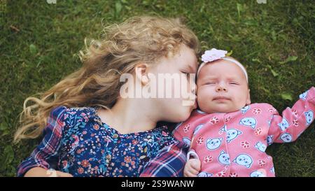 Blondes Mädchen küsst zärtlich neugeborene Geschwister, während sie sich auf grünem Gras ausruht, und fängt eine zärtliche familiäre Bindung und kindliche Unschuld ein Stockfoto