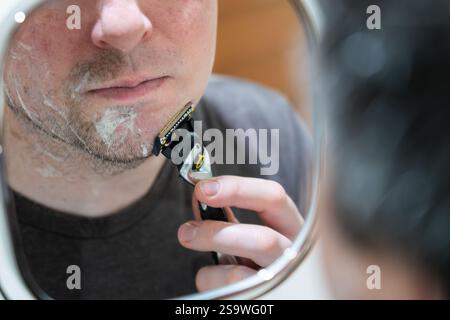 Mann mittleren Alters, der in einen wandmontierten ausziehbaren Spiegel schaut und sich mit einem Gillette Rasierer rasiert, Großbritannien. Konzept: Persönliche Pflege, morgendliche Routine Stockfoto