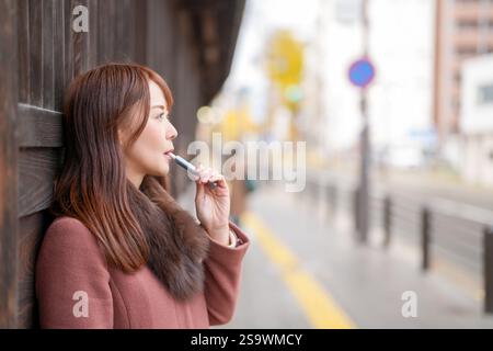 Neben den historischen Gebäuden in Japan. Eine Japanerin mit langen Haaren in den dreißiger Jahren schaut in die Stadt, indem sie elektronische Zigaretten raucht. Stockfoto