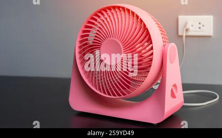 Nahaufnahme eines rosafarbenen elektrischen Tischventilators auf schwarzem Tisch mit weich verschwommener Rückseite des Steckers an der Wand. Selektiver Fokus. Stockfoto