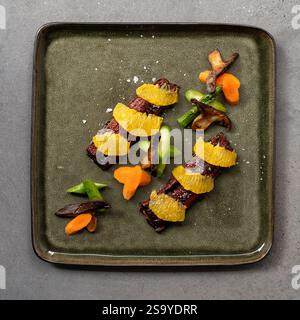 Blick von oben auf ein Gourmetgericht mit Orangenscheiben, gegrilltem Gemüse und Pilzen auf einer strukturierten grünen Platte. Stockfoto