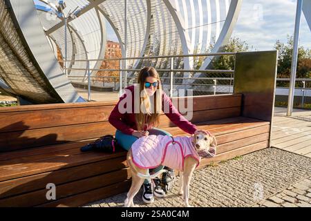 Junge Frau streichelt ihren windhund-Hund in einem rosa gesteppten Mantel, während sie auf einer Bank auf der arganzuela-Fußgängerbrücke in madrid sitzt Stockfoto