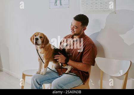 Porträt eines lächelnden Mannes, während er einen Beagle auf seinem Schoß hält, auf einem Holzstuhl in einem hell beleuchteten Raum sitzt Stockfoto